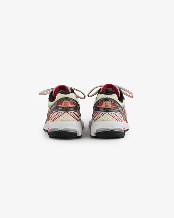 A pair of athletic shoes is displayed from the back, showcasing a beige and red color scheme with intricate detailing. The shoes feature a cushioned sole and prominent laces, designed for comfort and style.